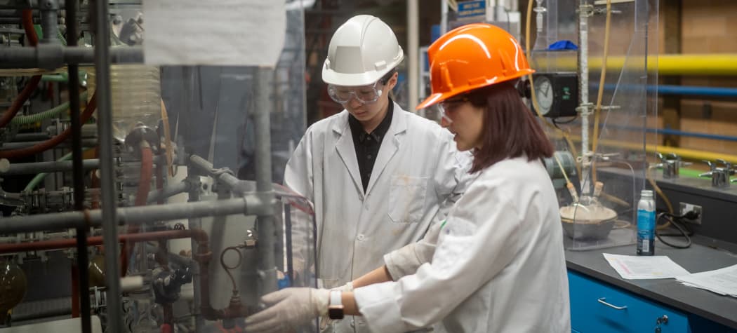 Two researchers wearing lab coats, hard hats, and safety goggles examine industrial laboratory equipment and piping.
