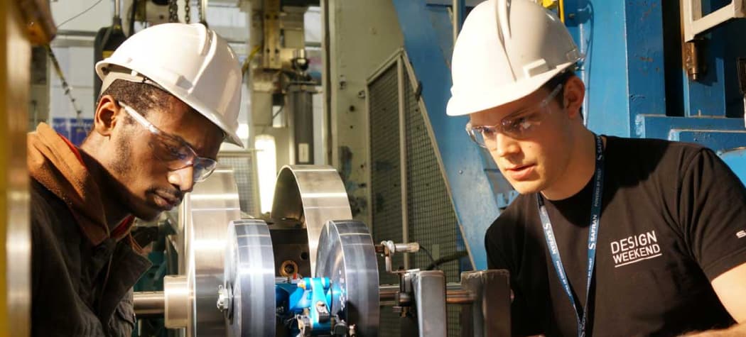 Two young male engineering students wearing white hard hats and safety glasses work closely together to inspect a mechanical component in a workshop.