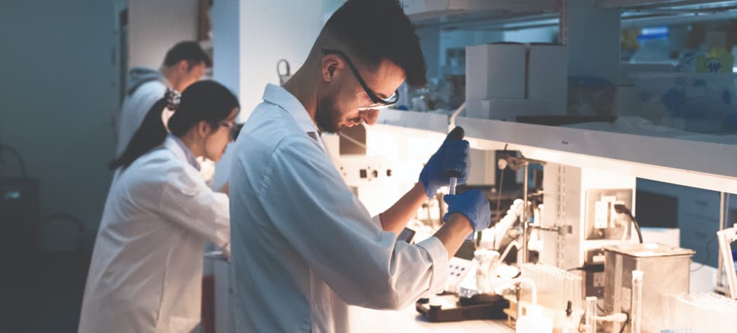 Scientists in white lab coats and blue gloves working in a modern research laboratory. One scientist in the foreground uses a pipette, while others work in the background.