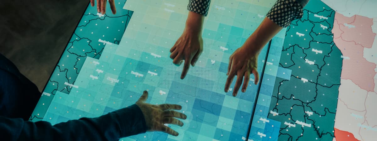 Three people point and gesture over a large illuminated digital map or data display table with shaded regions and outlined boundaries in blue and teal tones.