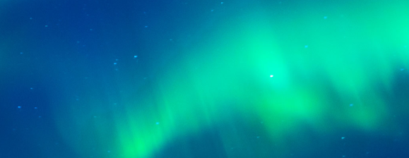 A long-exposure photograph of the Aurora Borealis (Northern Lights) shimmering across a dark night sky filled with faint stars. The aurora displays vibrant green and light teal hues, appearing as soft, ethereal curtains of light that wave vertically against a deep indigo and navy blue background.