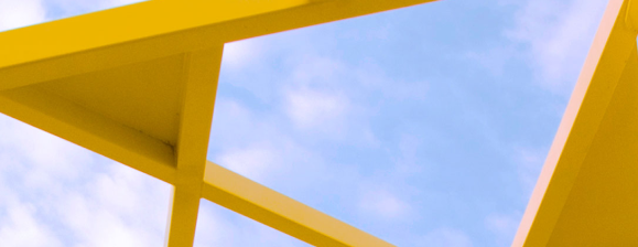 A minimalist, low-angle photograph featuring the sharp, geometric lines of a bright yellow structural frame or architectural installation. The yellow beams intersect to create triangular negative spaces, through which a pale blue sky with soft, wispy white clouds is visible.