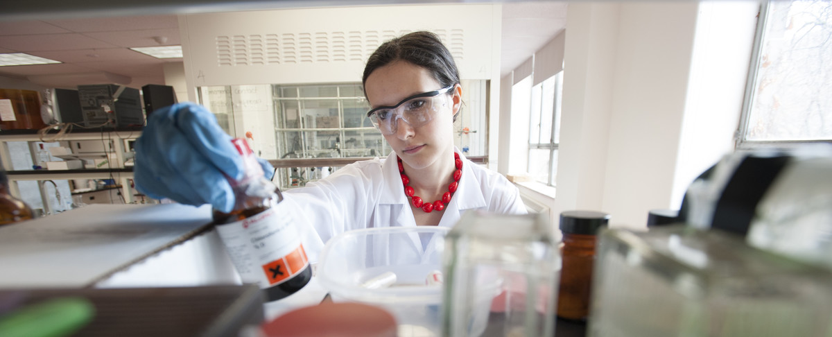 A scientist wearing safety glasses and blue gloves works in a laboratory, reaching for a chemical bottle among glass containers and lab equipment. Bright windows and shelves of scientific instruments are visible in the background.