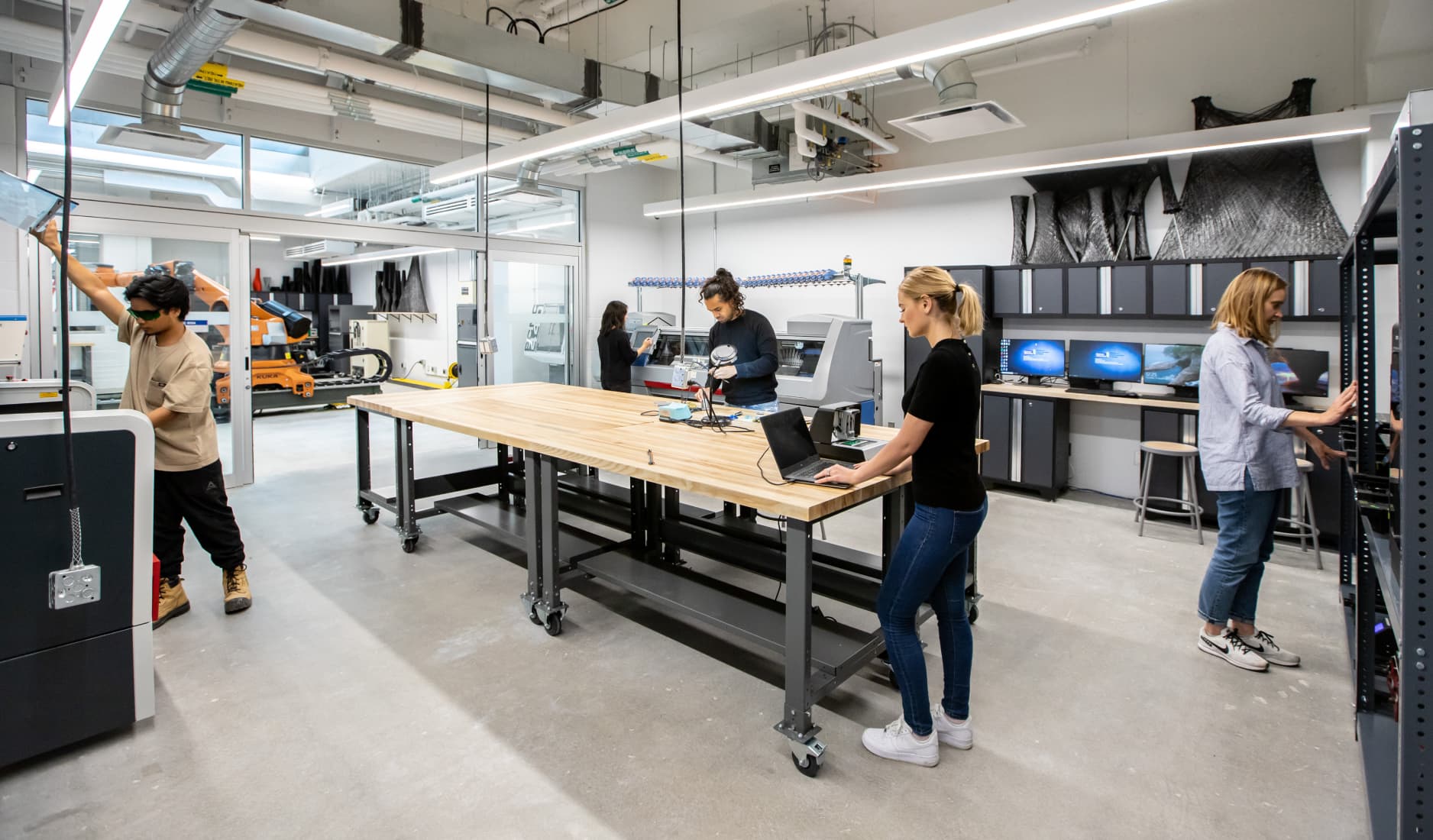 Students working in a modern, well-equipped maker space or engineering lab featuring large wooden workbenches, 3D printing equipment, and computer workstations.