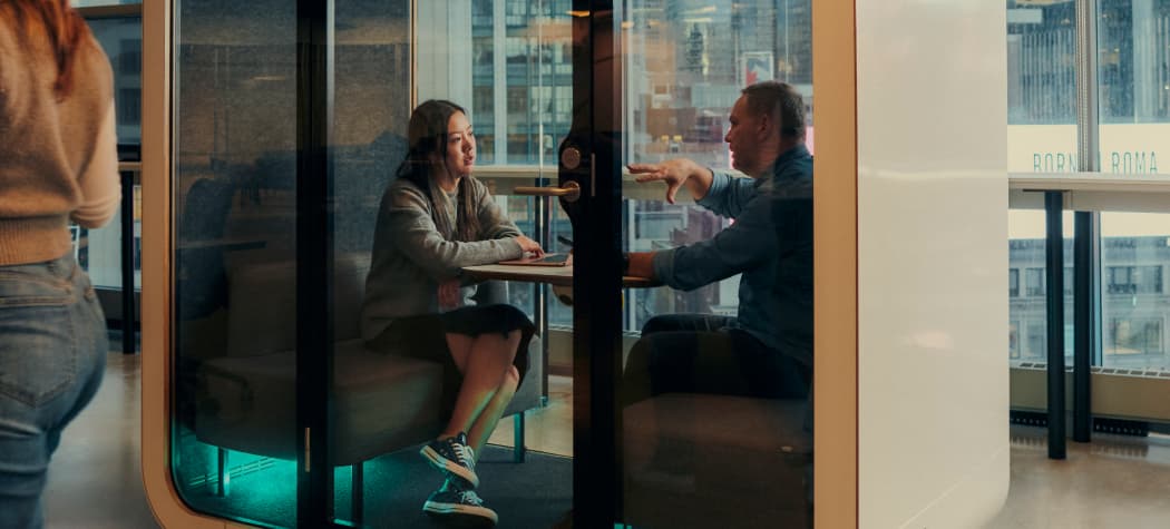 Two people sit facing each other in a glass-walled office pod, engaged in conversation. The woman wears a blazer and sneakers, while the man gestures with his hands. A blurred person walks past in the foreground, and city buildings are visible through the windows in the background.
