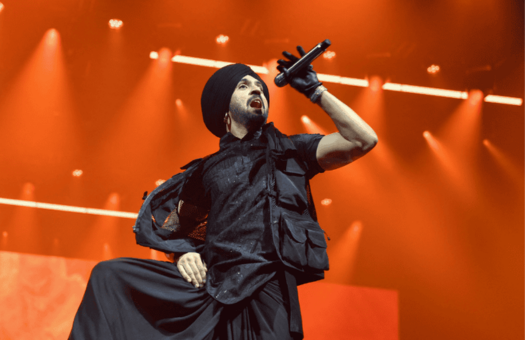 Diljit Dosanjh singing into a microphone on stage under bright orange concert lighting.