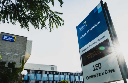 Outdoor view of Toronto Metropolitan University’s School of Medicine signage at 150 Central Park Drive, with campus buildings in the background, trees in the foreground, and sunlight flaring near the sign.