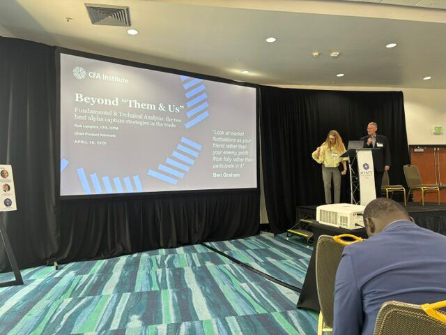A conference room with a large presentation screen displaying "Beyond 'Them & Us'" by CFA Institute. Two speakers are at a podium.