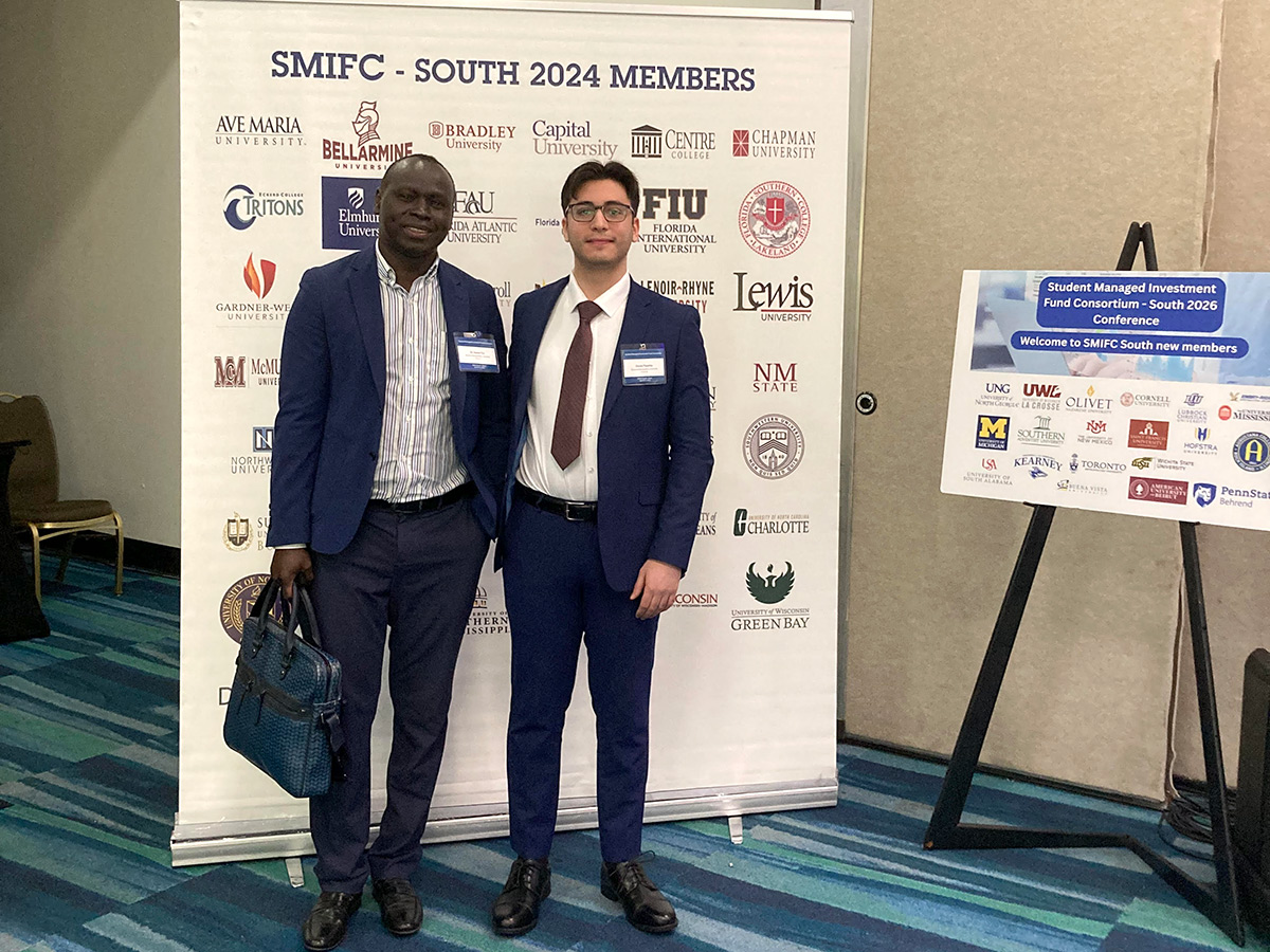 Professor Daniel Tut and student Daniel Panatt stand smiling in front of a backdrop displaying university logos at the SMIFC South 2026 event.