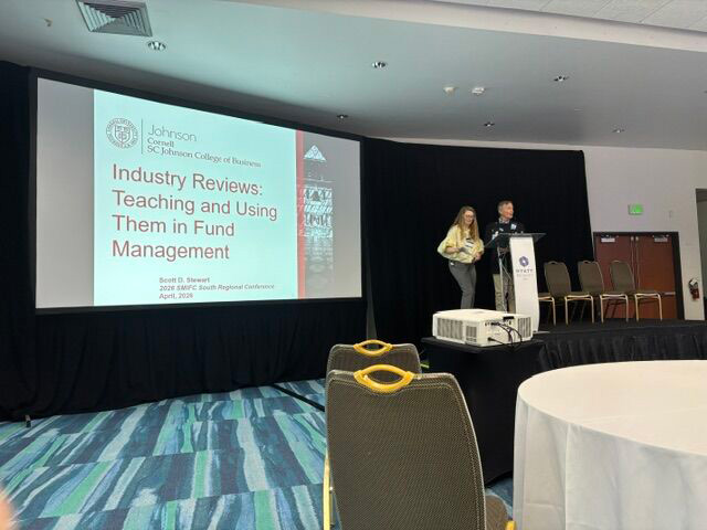 A conference room with two presenters at a podium beside a large screen displaying a presentation titled "Industry Reviews: Teaching and Using Them in Fund Management."