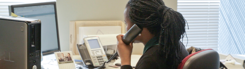 A side view of a woman at an office workstation, facing her computer screen and speaking on the phone.