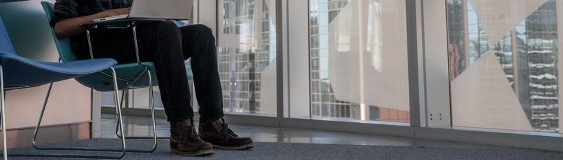 View of seated man from arms extended to laptop down to his feet sitting flat on the floor with the Student Learning Centre windows in the background.