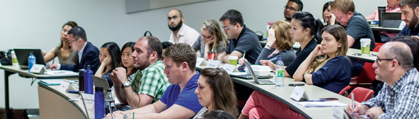 Lecture-style classroom filled with students looking towards the front of the room, using paper and laptops to take notes.