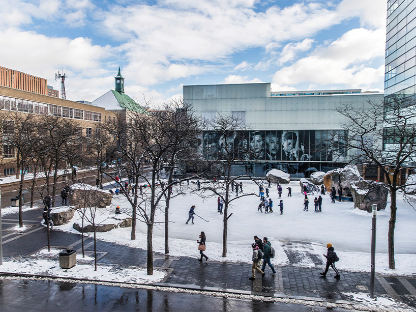 Students walk along the intersection of Victoria Street and Gould Street, while others skate on a frozen Lake Devo on a bright winter day.
