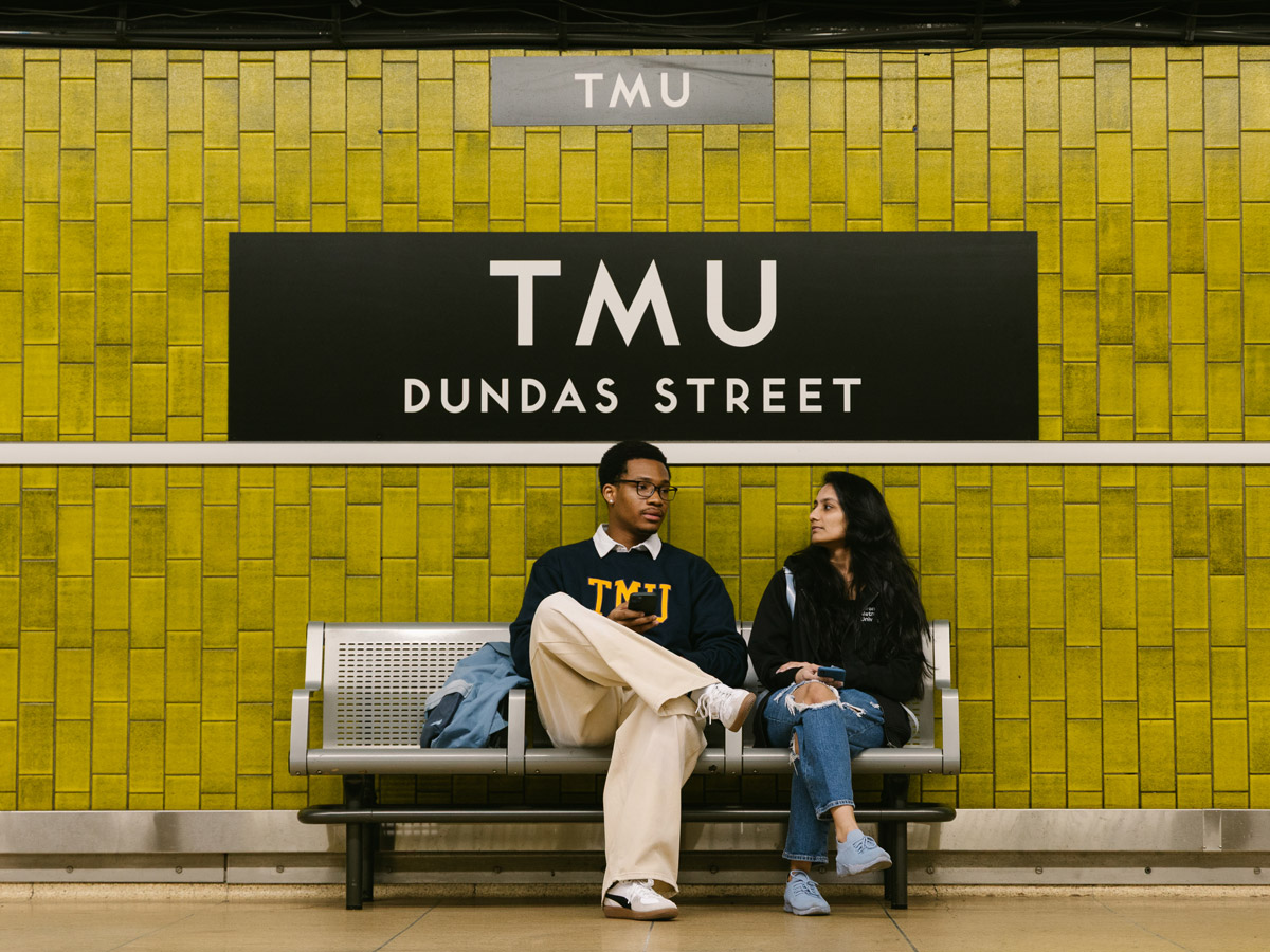 A pair of students sit on a bench under the TTC sign for TMU Station.