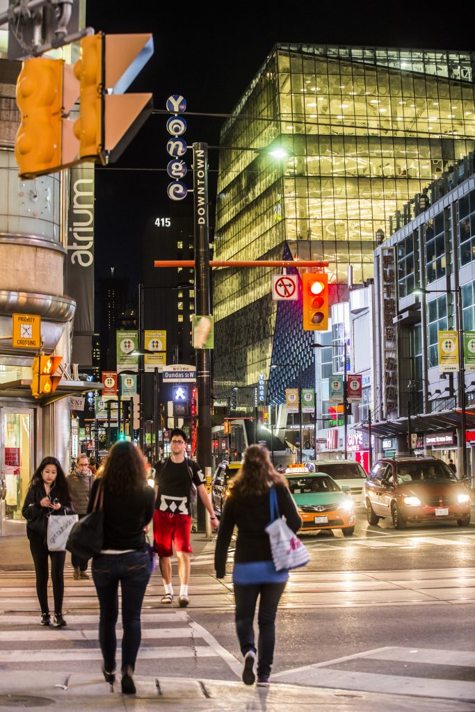 Pedestrians crossing the street near Yonge-Dundas square and can see the Student Learning Centre in the background in the night.