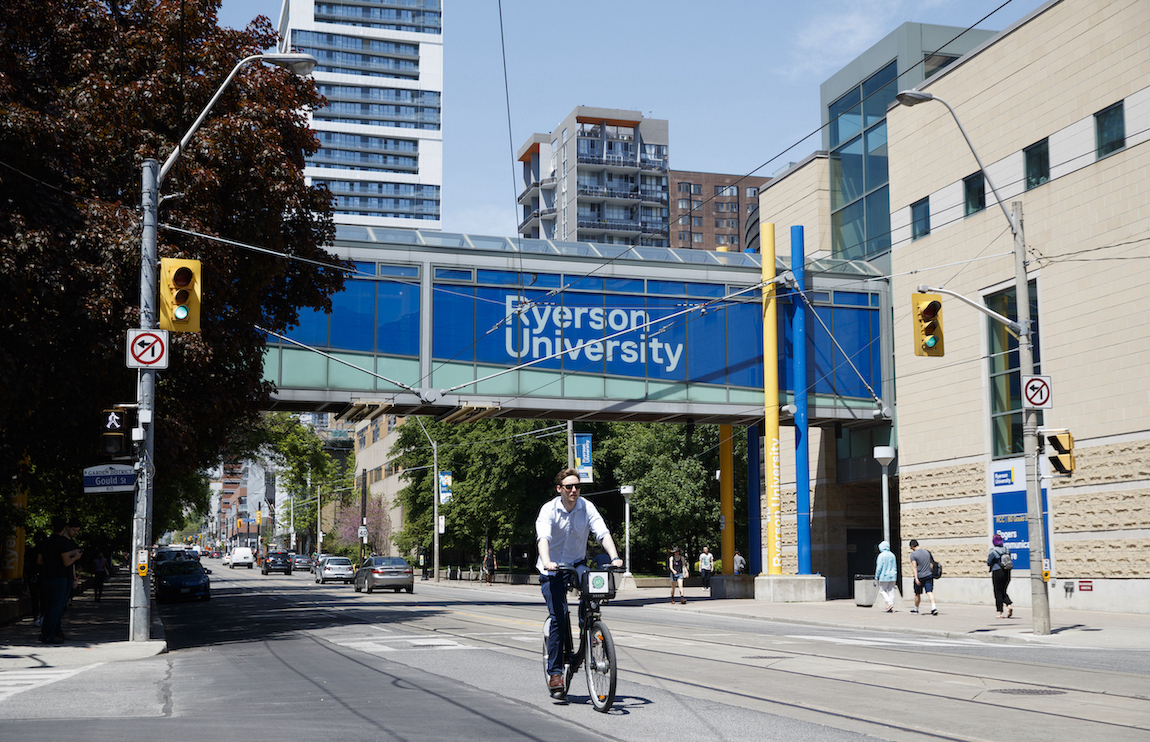 Sunny day, person riding bike past the Ryerson birdge that connects Kerr Hall and Rogers Communications Centre building