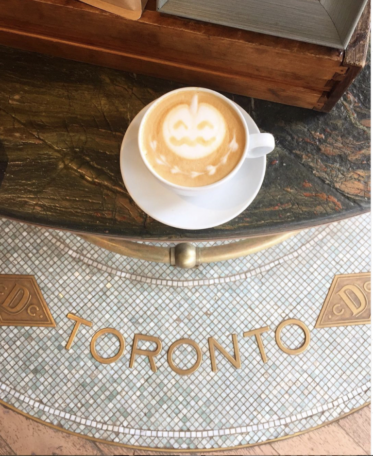 Pumpkin art in a cup of coffee, with the letters Toronto on the floor tiling in a clean and rustic design