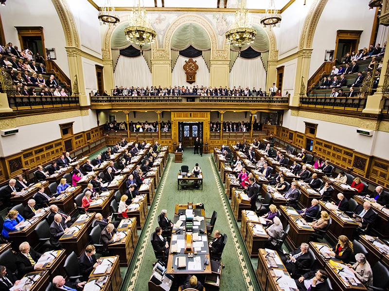 A session of government is in session in a government buildings with members of parliament sitting at their desks and onlookers watching from the galleries above.