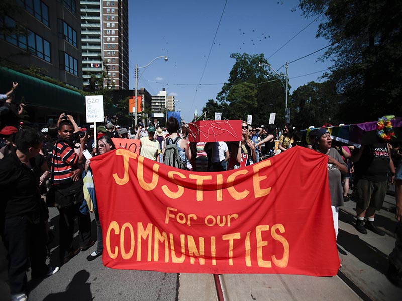A group of people protest while walking down the street and holding a large red banner with  "Justice for our communities" written in yellow.