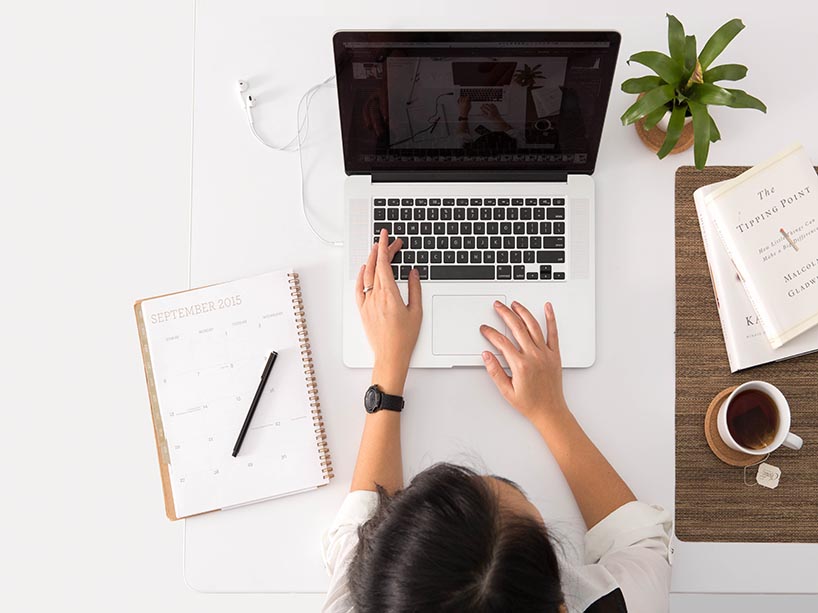 A birds-eye view of a woman working on her computer on a white desk. On the white desk is a notebook planner, white headphones, a plant, and a cup of tea. 