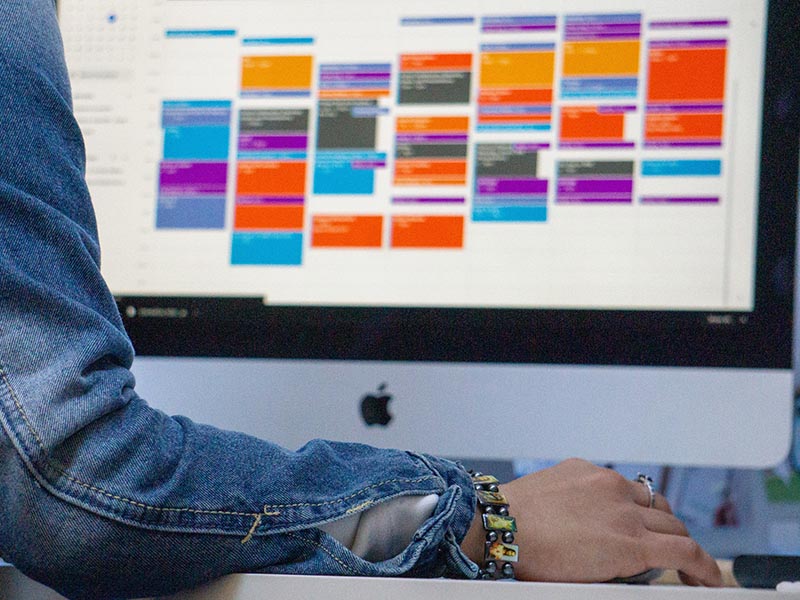 A woman sits in front of her desktop wearing a jean jacket and chunky jewelry. On her desktop is a cluttered and crowded Google Calendar.