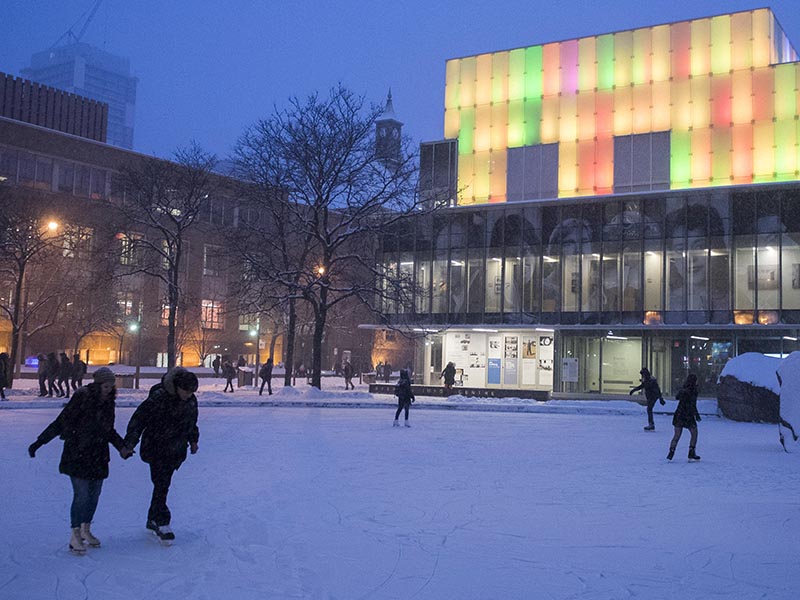 A couple skates while holding hands in the blue light of early nightfall on our campus' Lake Devo with the Image Centre lit up in the background against the dark sky. 