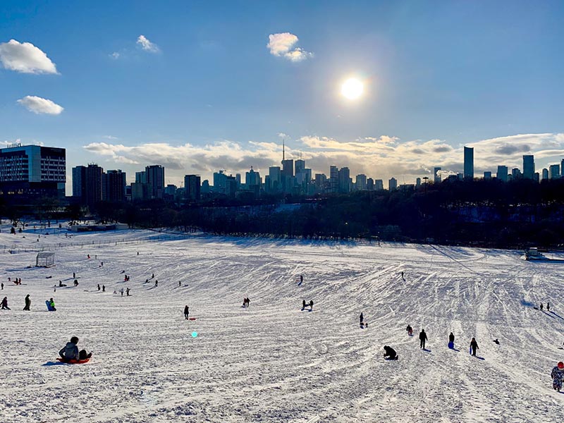 Riverdale Park East on a bright sunny afternoon. Children climb and slide down the snowy hill with Toronto's city skyline in the background. 