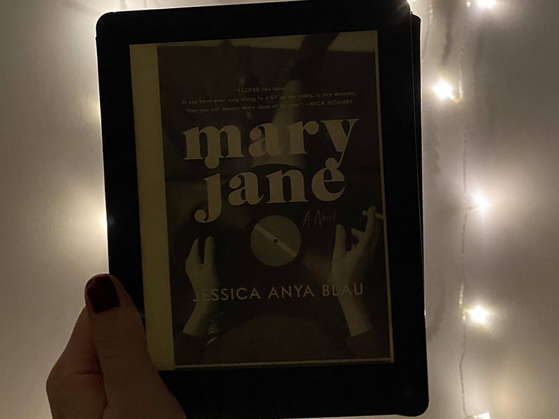 The cover of the book Mary Jane by Jessica Anya Bleu on my ereader in front of a background of a white wall and twinkle lights. The book cover is of two hands holding a record.