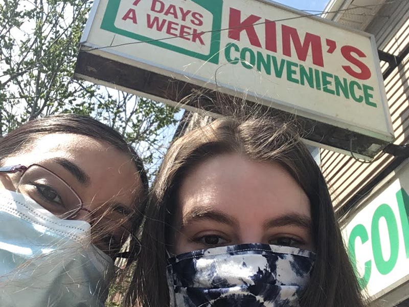 Jenna and Melissa standing under the Kim's Convenience storefront while wearing masks on a bright, sunny day.