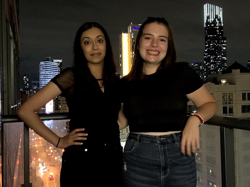 Jenna and Melissa smile on a balcony at night time with bright city lights of buildings in the background.