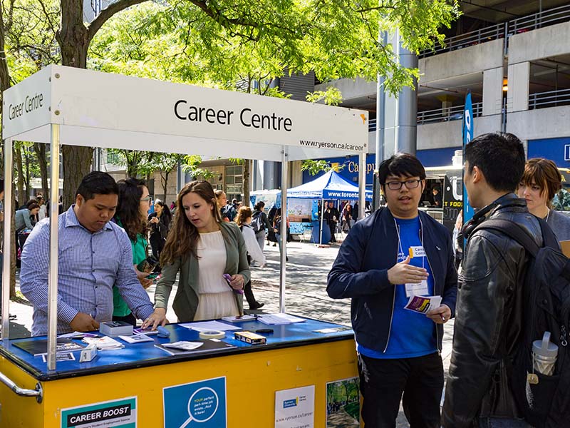 Workers for the Career Centre stand behind and in front of their booth, talking to students during a sunny afternoon on Gould Street.