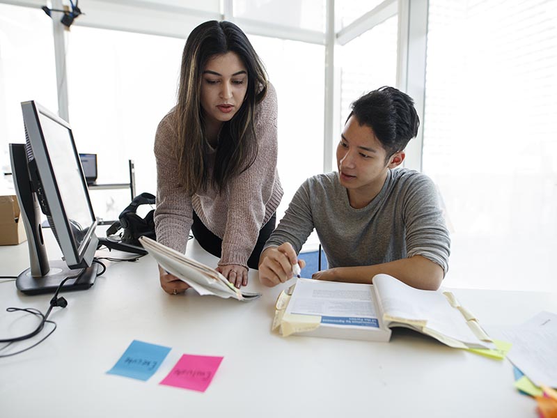 Two students work together while looking at a textbook and a notebook.