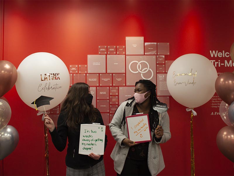 Two women pose smile and laugh at a Tri-Mentoring Program celebration event while holding whiteboards with their goals.