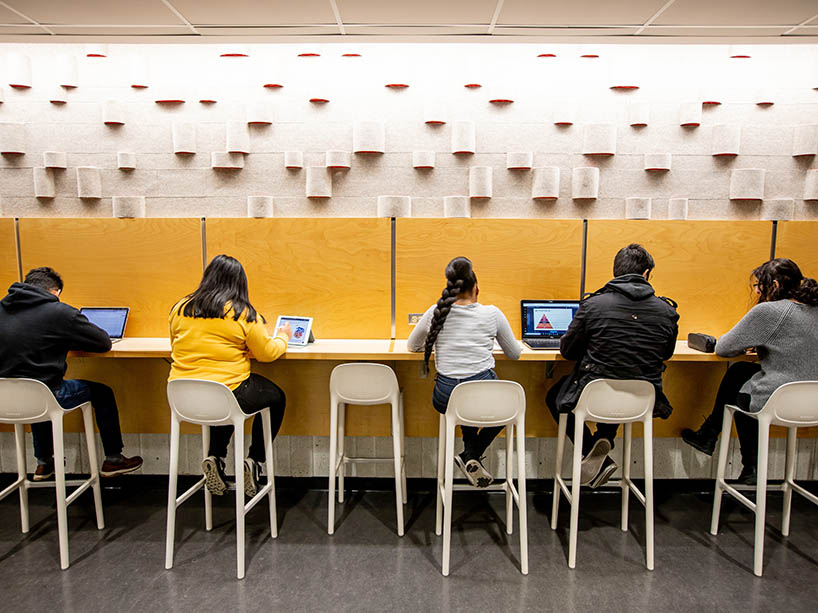 Students work on their computers and tablets on high stools facing a wall.