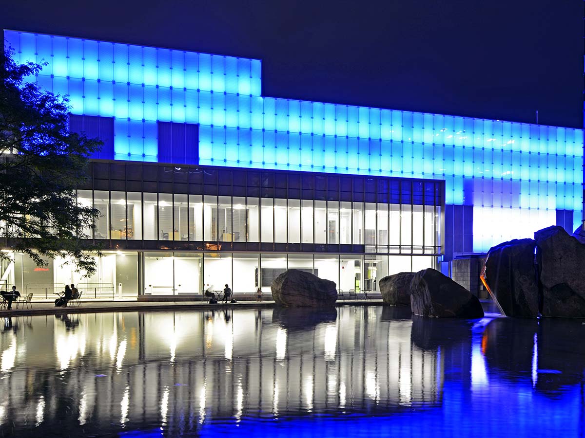 The exterior of the Image Centre lit up in light blue against a night sky, with the blue reflecting in Lake Devo below.