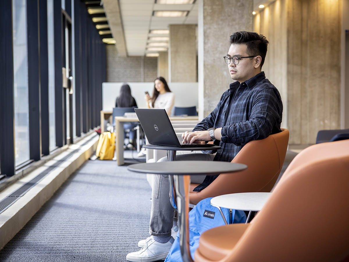 A student works on his laptop in the library in a chair with a laptop stand, facing the exterior windows.