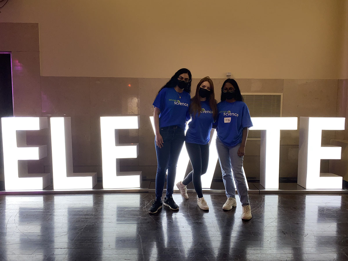 Lynn and her friends in front of a lit-up sign that reads "ELEVATE".