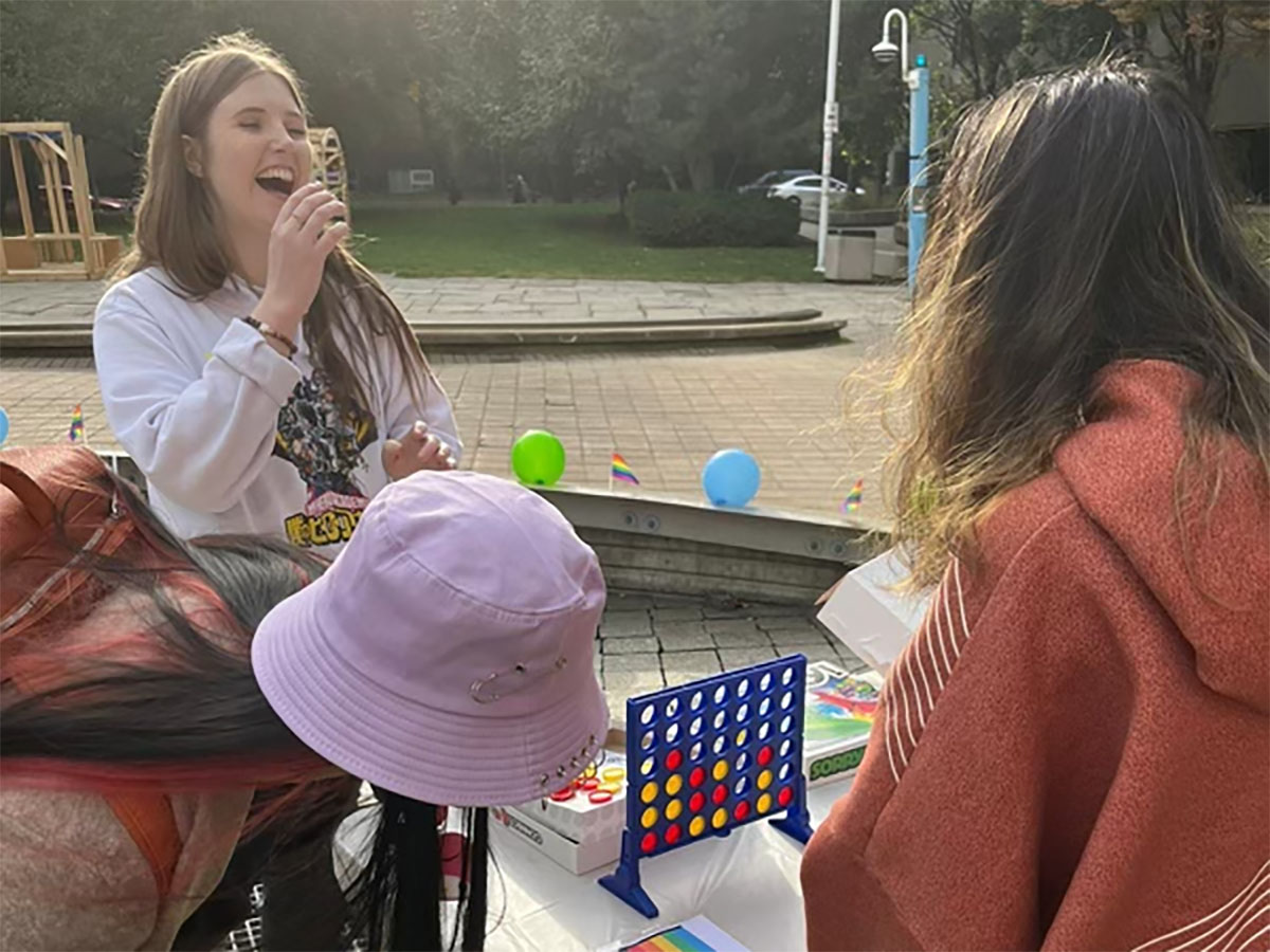 Sophie laughs and plays a game of Connect Four in Pitman Quad during the Ted Rogers Pride Alliance queer mixer on a sunny afternoon.