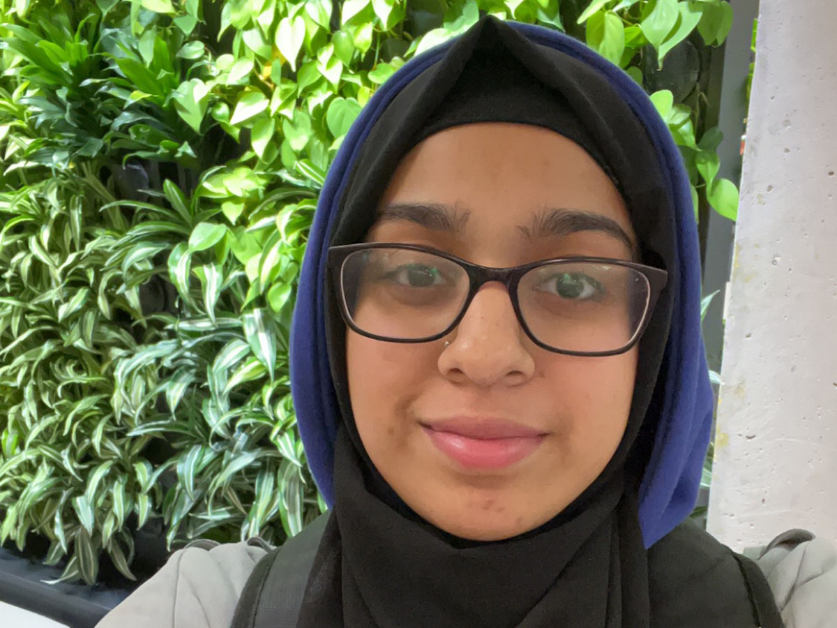 Hooria takes a selfie in front of a wall of green plants.