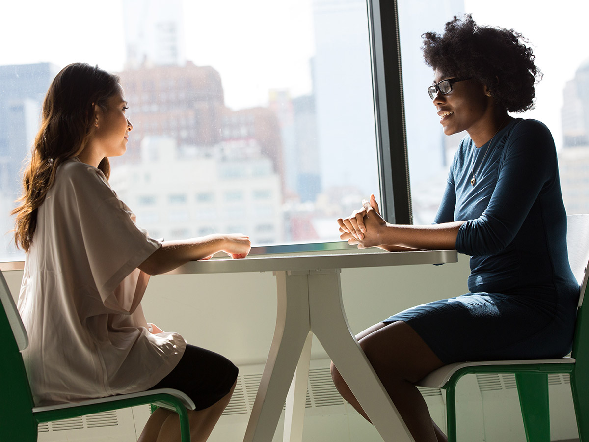Two women talk at a small round table in front of large windows.