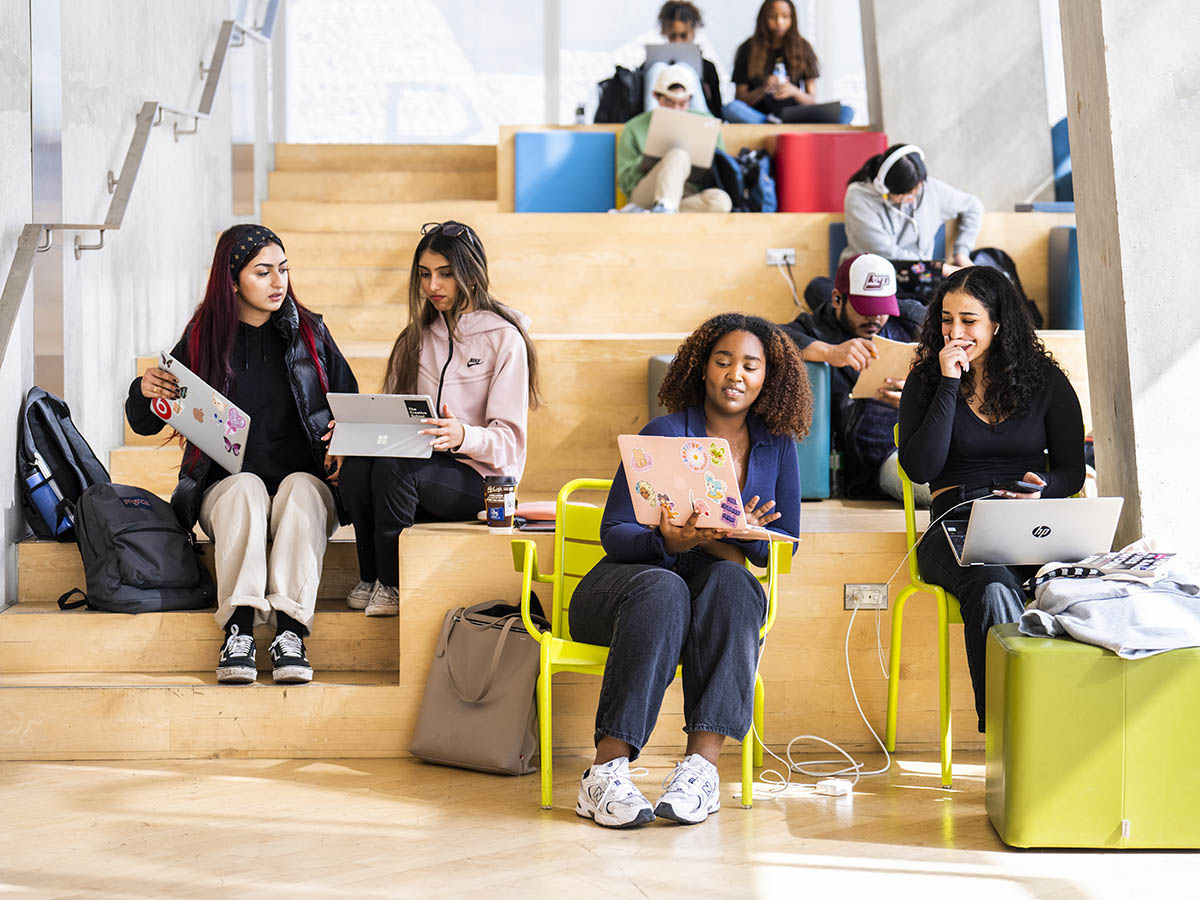 TMU students sit on wooden steps and chat with each other while on their laptops.