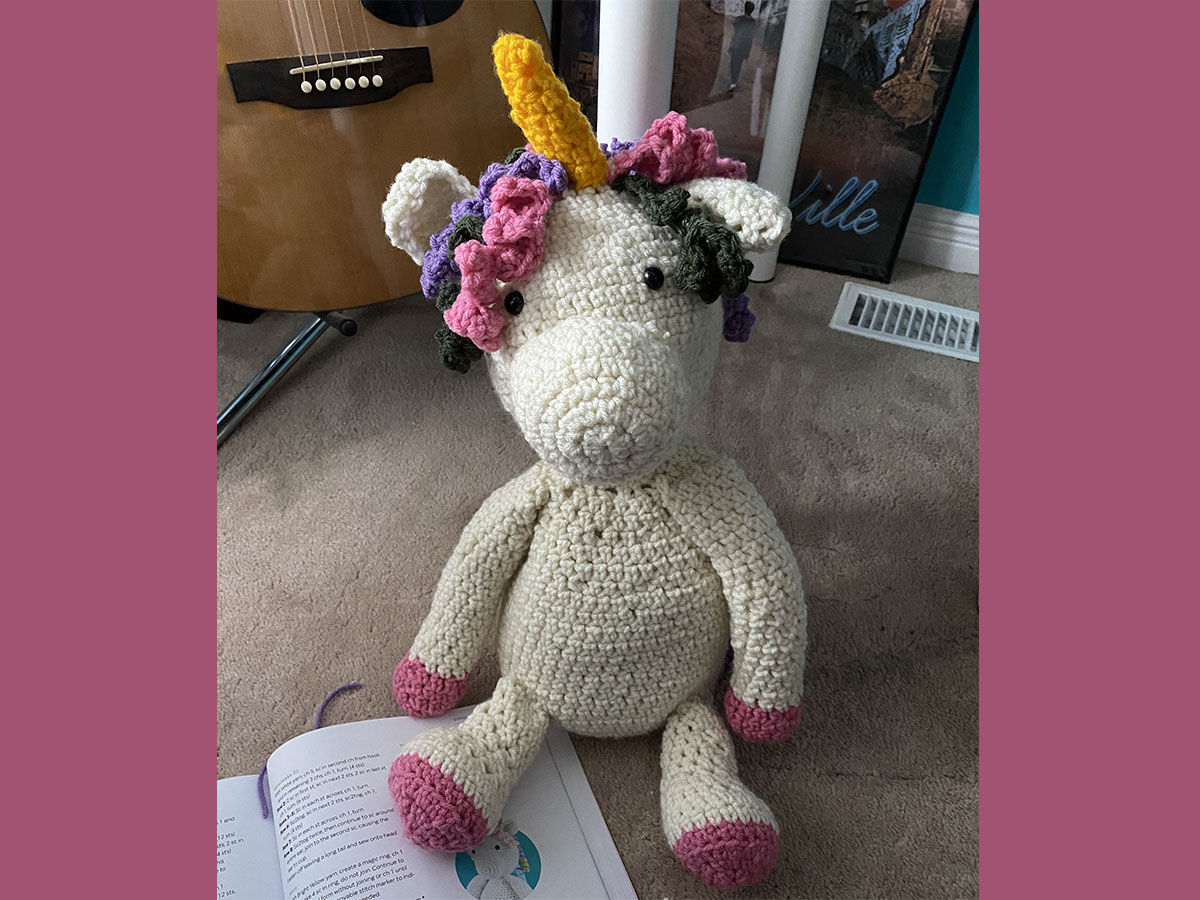 A white crocheted unicorn with pink paws, a yellow horn and colourful hair.