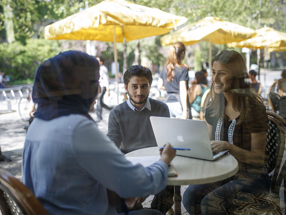 Three students huddle around a cafe table studying on campus. There is a laptop and notebook on the table.