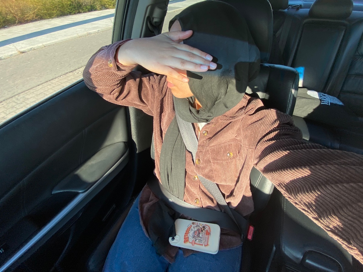 Hadiqah is sitting in a car with a hand over her face, wearing a brown shirt.