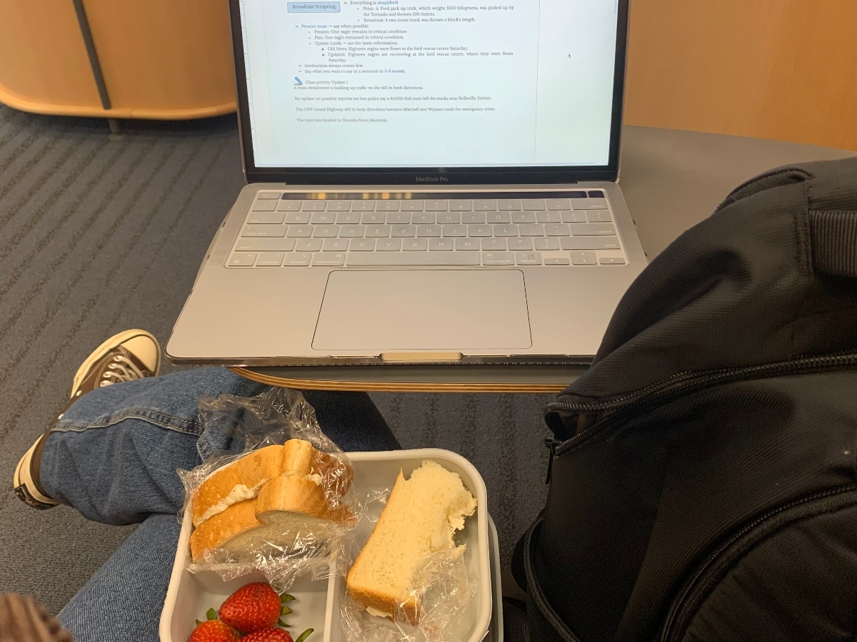 A lunch box with strawberries and a sandwich in Hadiqah's lap. A laptop is on the table above her.