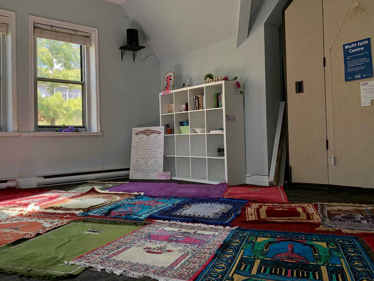Sign that says "multi-faith centre" on the right and the ground is covered in prayer mats as sunlight falls onto them from windows on the left.