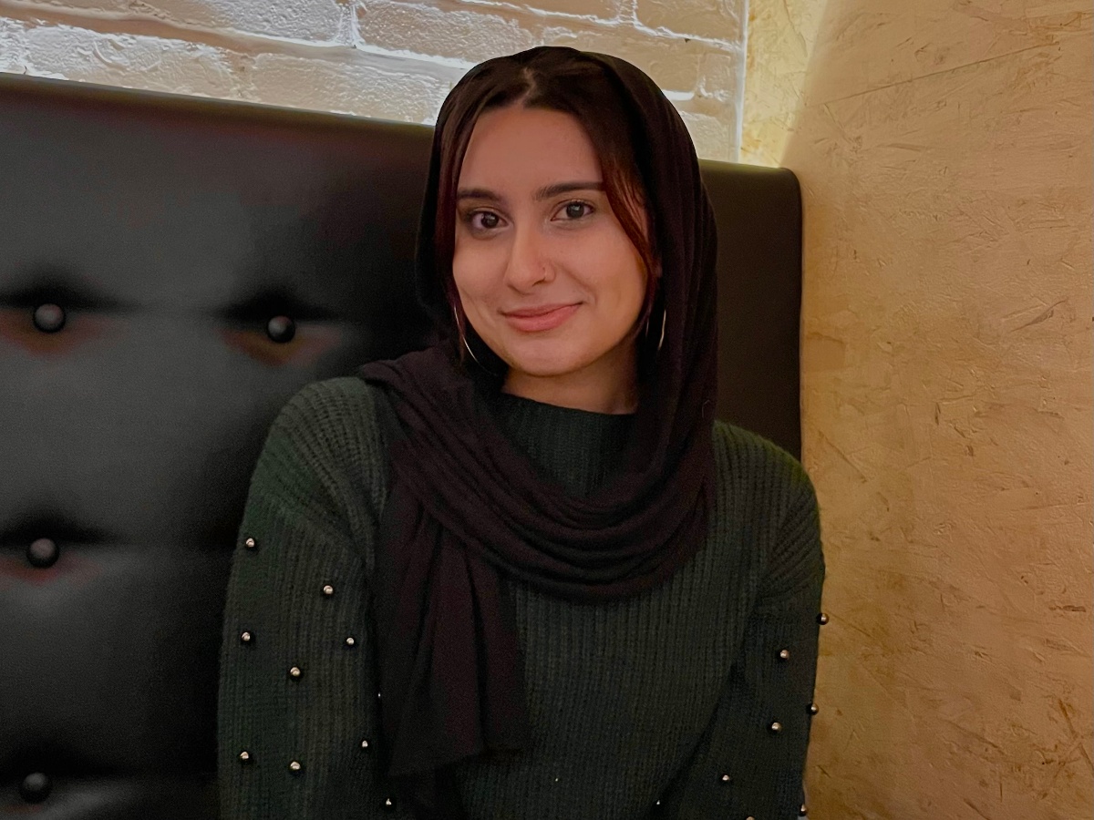 Fiza is smiling wearing a black hijab wrapped around her head and a dark green shirt.