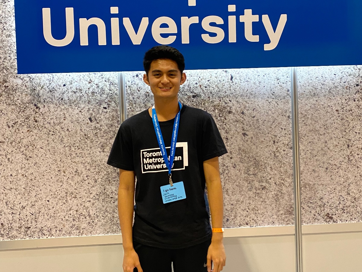 Jack is wearing a shirt that says Toronto Metropolitan University and has a lanyard across his neck. He is standing in front of a sign of the TMU logo and smiling.