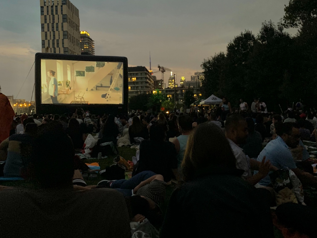 A projector screen showing a film outdoors, as people are sitting on the grass in blankets while the sun sets.